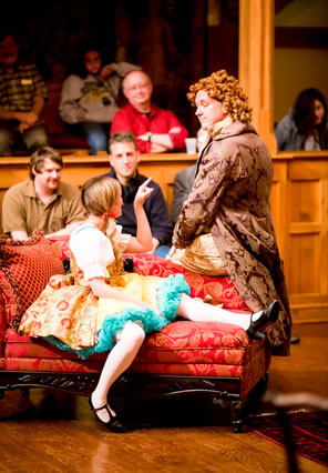 Prue, in yellow and white country dress with frilly blue trim and white stockings sits spread-legged on chaize lounge pointing up at  Tattle in gold brocade coat and curly orange wig sits on the lounge back, with audience in the background.
